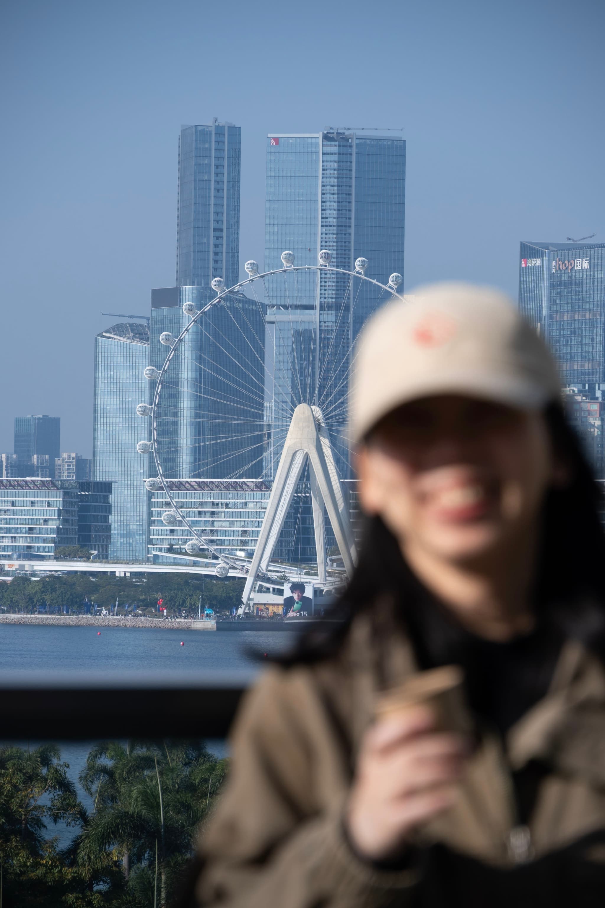 Out of focus person in a cap in the foreground with a ferris wheel and modern city skyscrapers in the background across the water