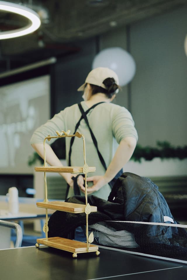 Baker in apron and cap working at a table with a pastry stand in a cafe or bakery setting