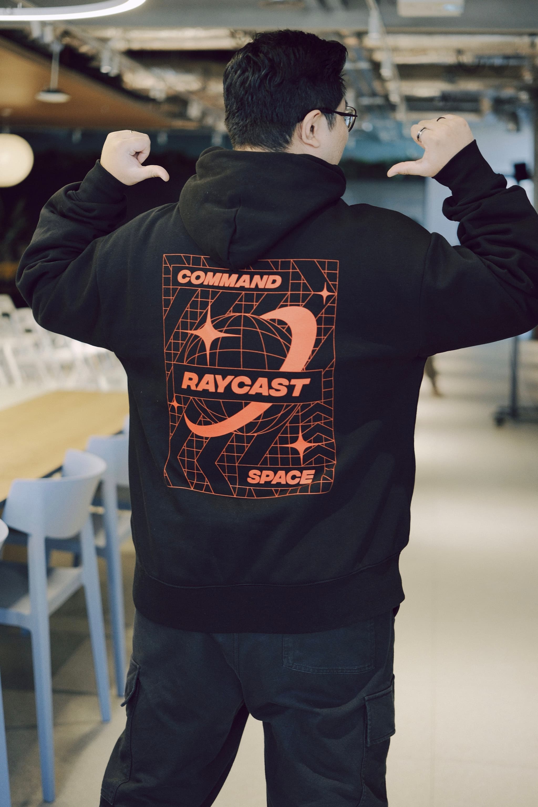 Person posing in a black hoodie with a bold orange graphic on the back reading Command Raycast Space in an indoor setting