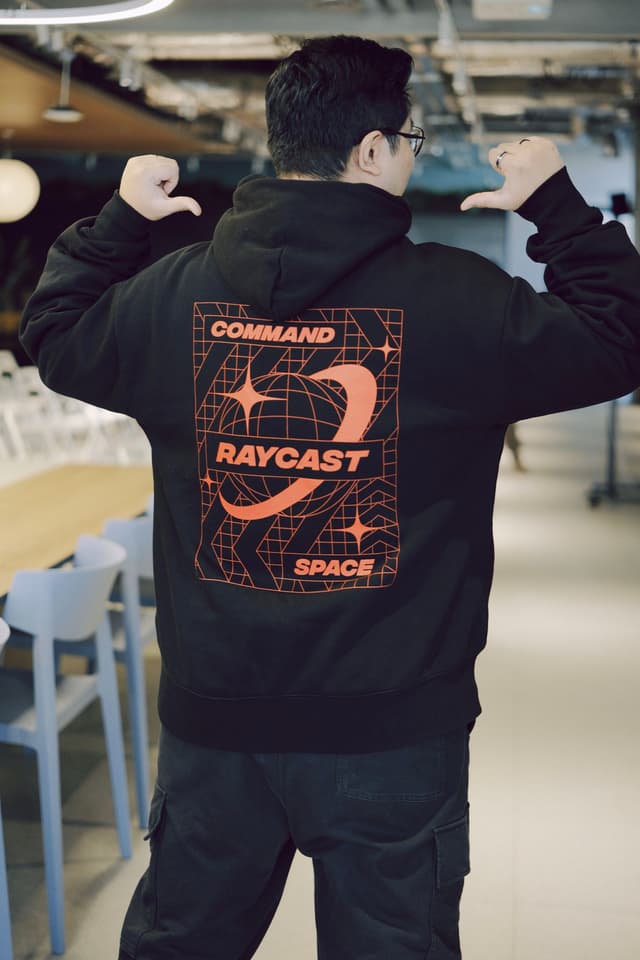 Person posing in a black hoodie with a bold orange graphic on the back reading Command Raycast Space in an indoor setting