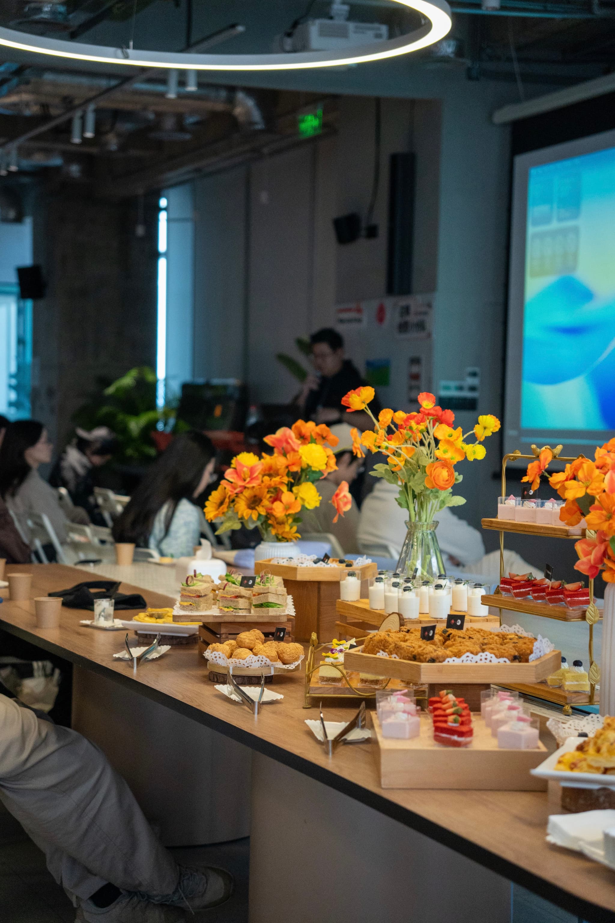 Modern conference room with people seated at long tables and a buffet spread with flowers while a presentation is projected on a screen