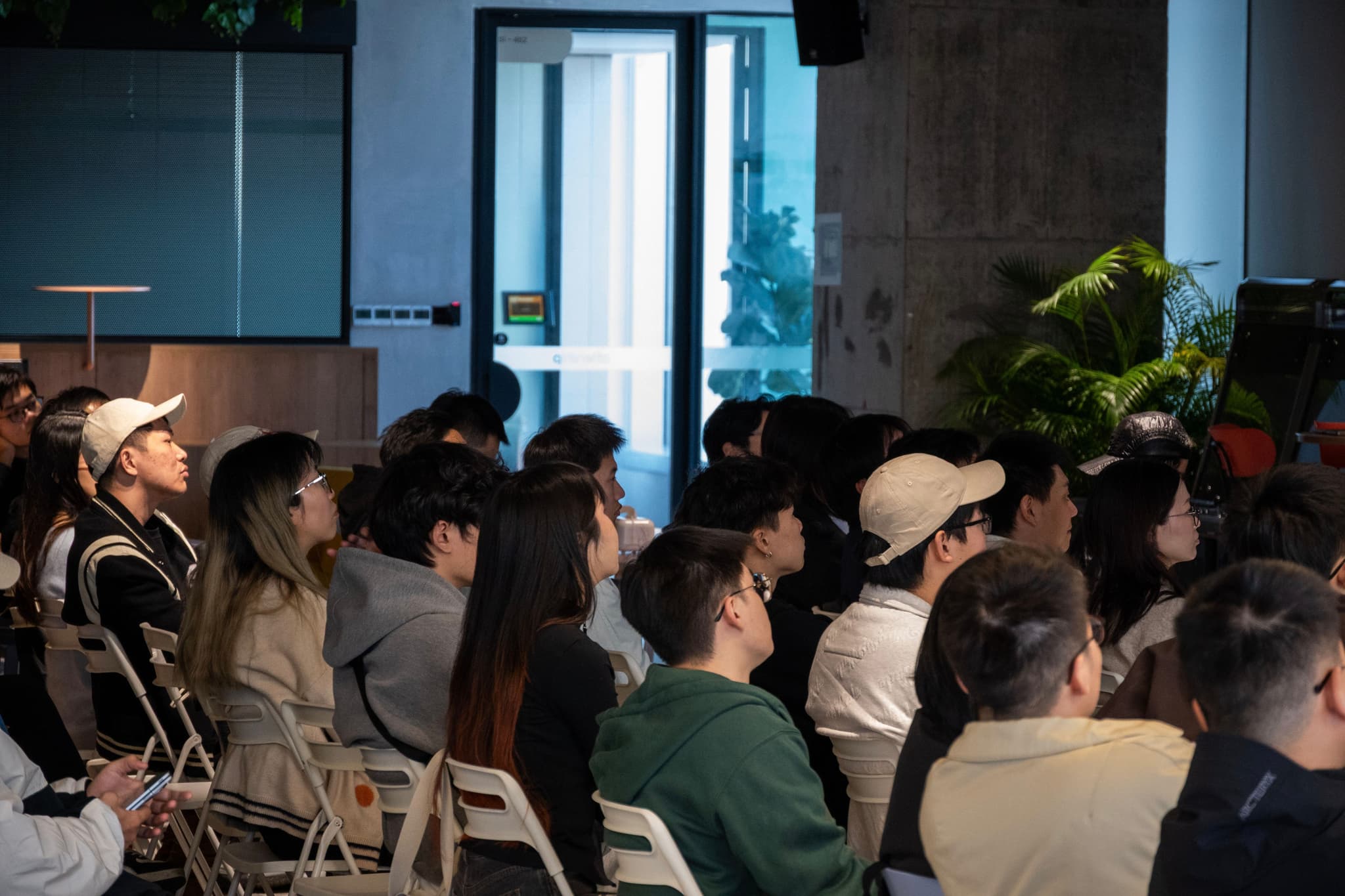 Side view of a seated audience in a dimly lit room facing a presentation area near a doorway with plants