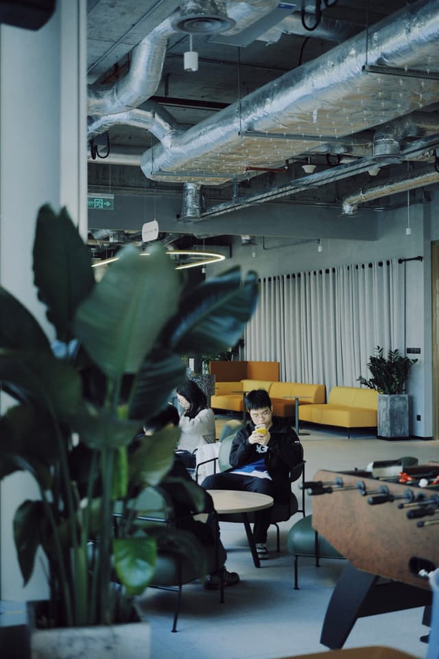 Industrial style office with exposed ductwork indoor plants and a person working at a small round table near a lounge area