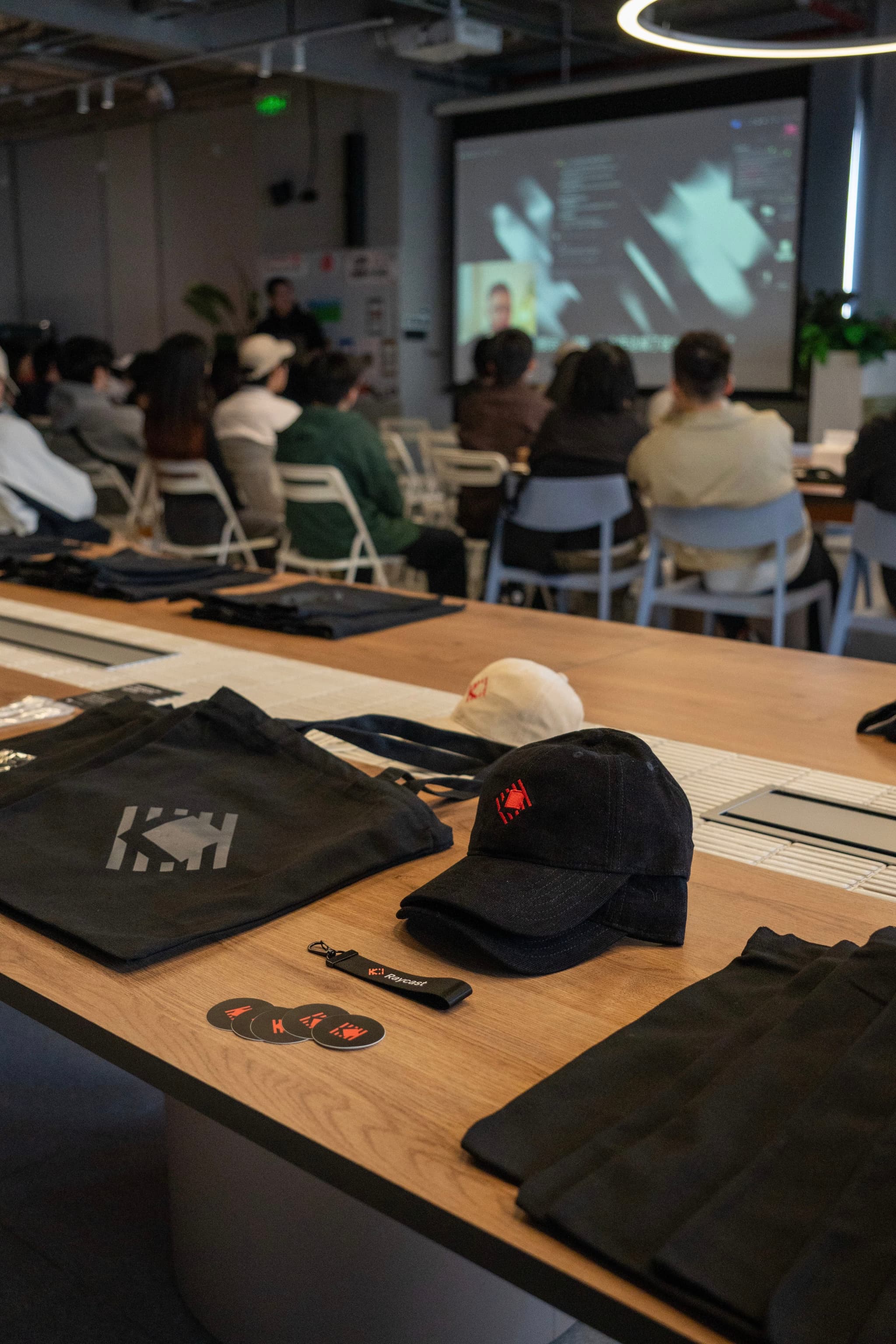 Branded merchandise on a table in foreground with an audience seated in a modern room watching a projected presentation