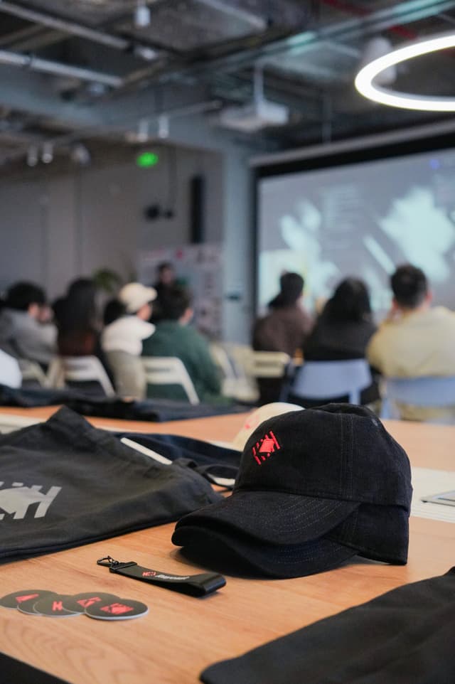 Baseball cap and tote bag on a table in the foreground with an audience watching a presentation in a contemporary conference room