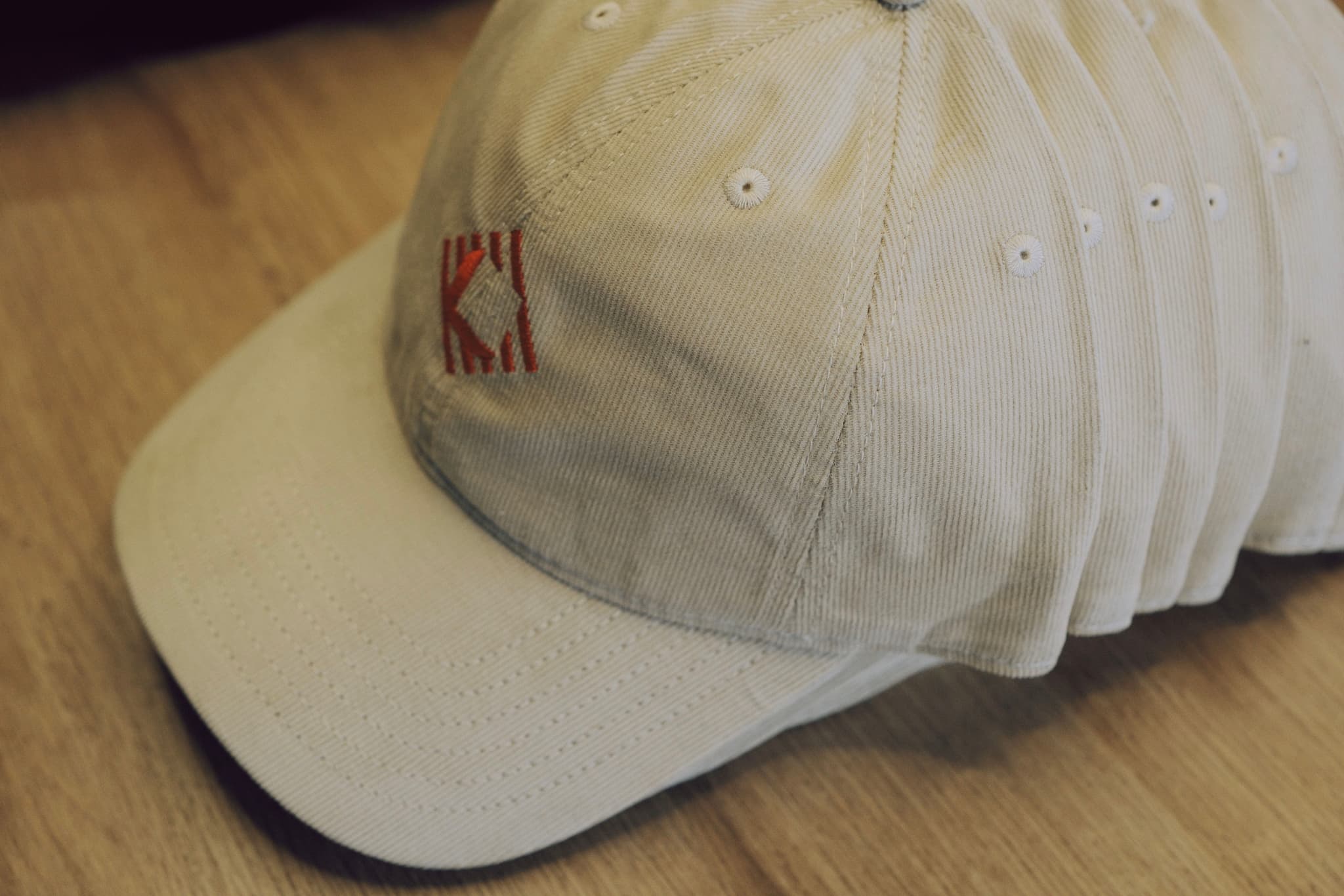 A stack of light colored baseball caps with embroidered logos on a wooden surface