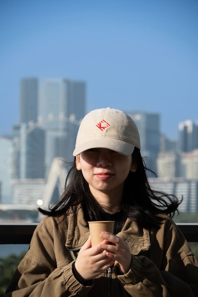 Person in a beige cap holding a coffee cup outdoors with a blurred city skyline in the background