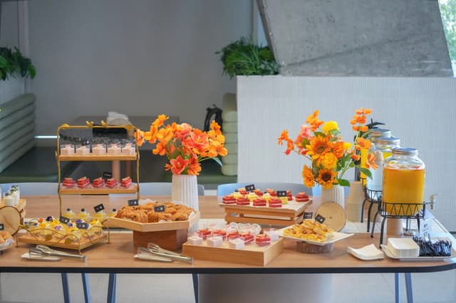 Indoor buffet table with assorted pastries desserts and drinks decorated with orange and yellow flowers against a modern gray wall