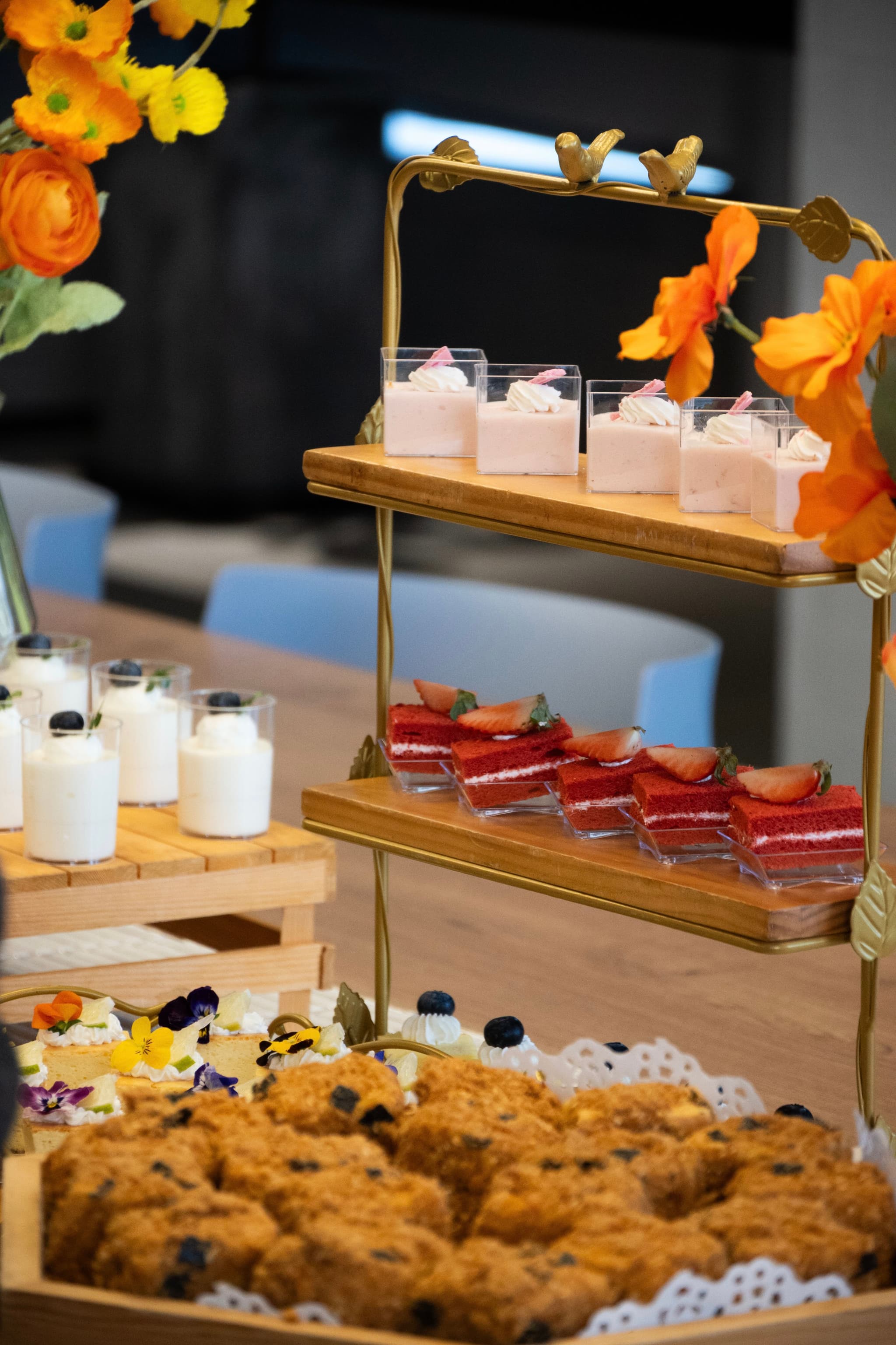 Tiered gold stand with tea cups and red drinks beside assorted pastries and cookies on a table with orange flowers