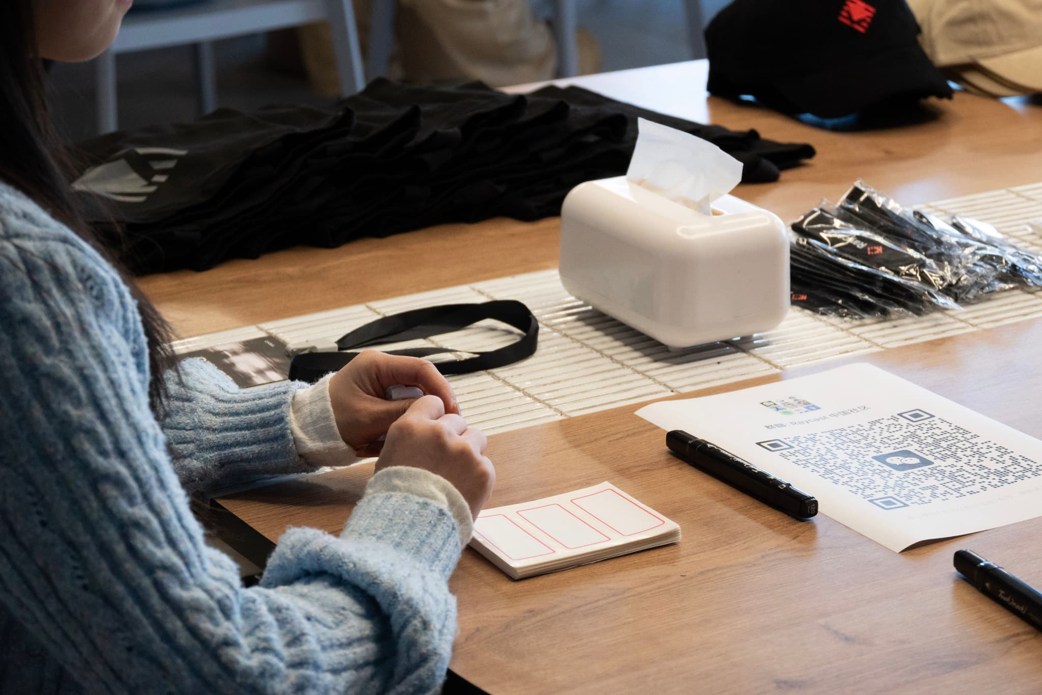 Person in a sweater sketching on paper at a wooden desk with a smartphone a black pen scattered documents and a small white device