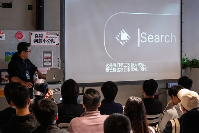 Presenter speaks to a seated audience in a conference room while a projector displays a search interface on a screen