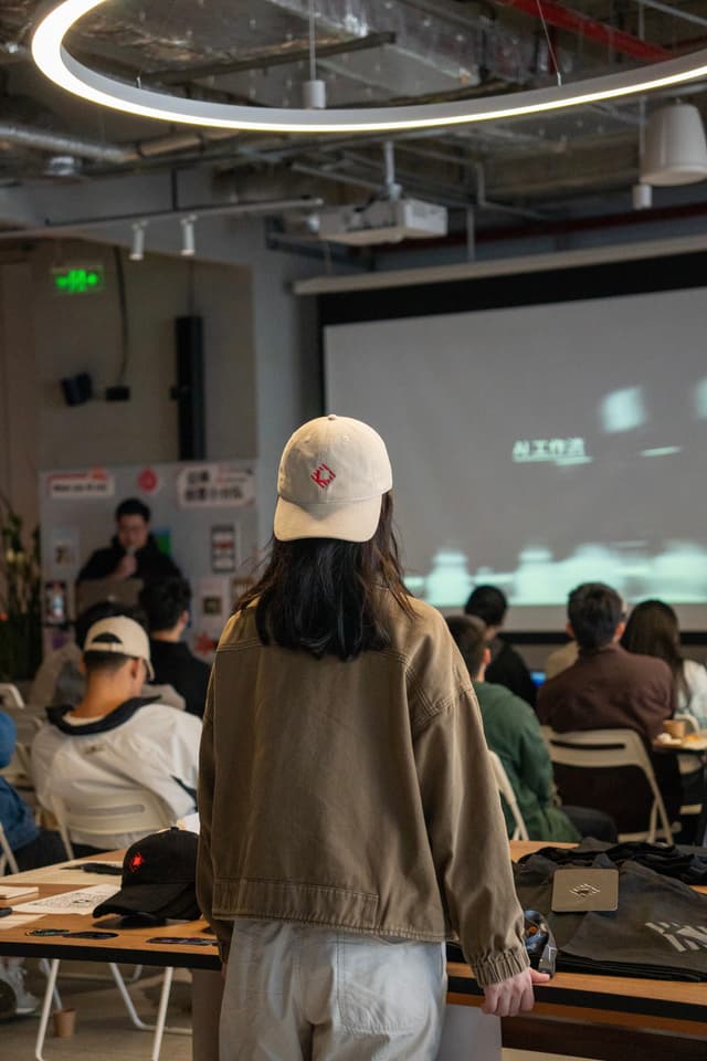 Person in white cap stands in a modern co working space facing a projected screen while seated attendees watch