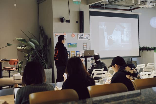 Small meetup room with attendees seated on couches facing a projected presentation screen and a speaker at the front