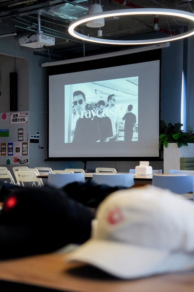Modern workspace with tables and hats in foreground and a large projector screen showing a group photo under circular ceiling lights