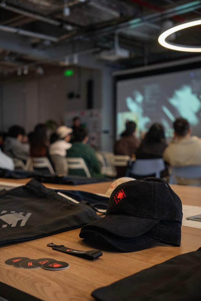 Caps and tote bag on a table in a conference room with a blurred audience watching a presentation on a screen