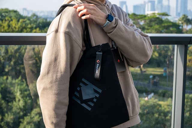Person in beige jacket holding a black tote bag on an outdoor balcony with a city skyline in the background