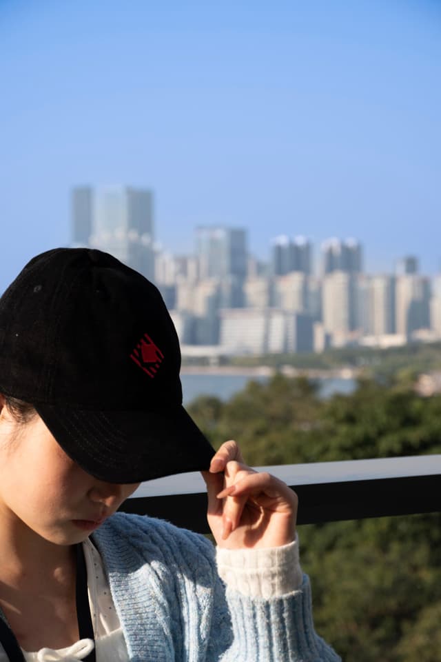 Person in a black cap adjusting the brim on a balcony with a blurred city skyline and blue sky in the background