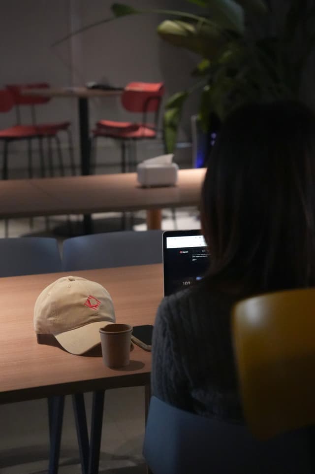 Person seated at a café table with an open laptop, coffee cup, and baseball cap, with empty chairs in the background