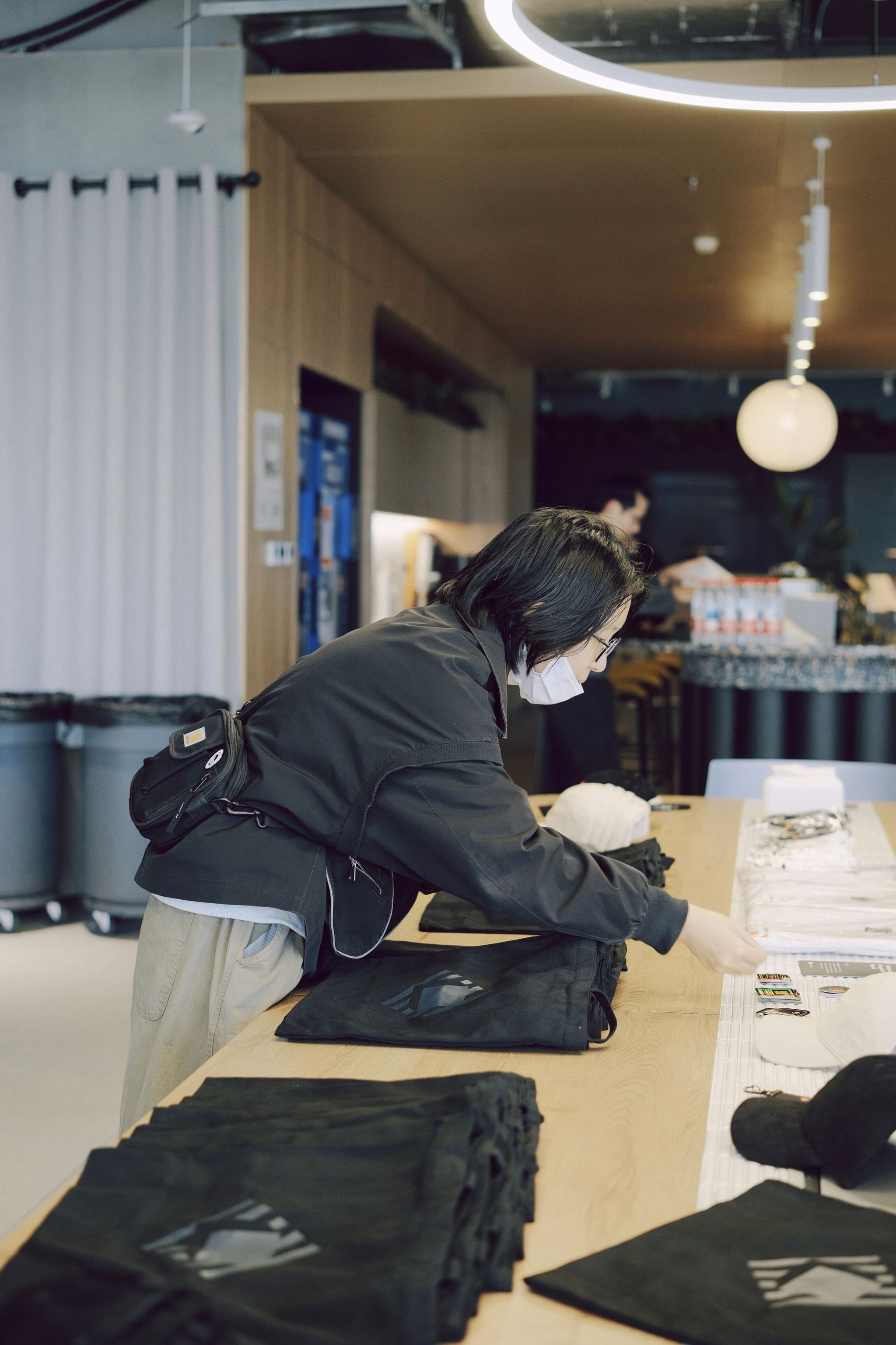 Person leaning over a tote bag at a counter with bags and clothing items laid out nearby