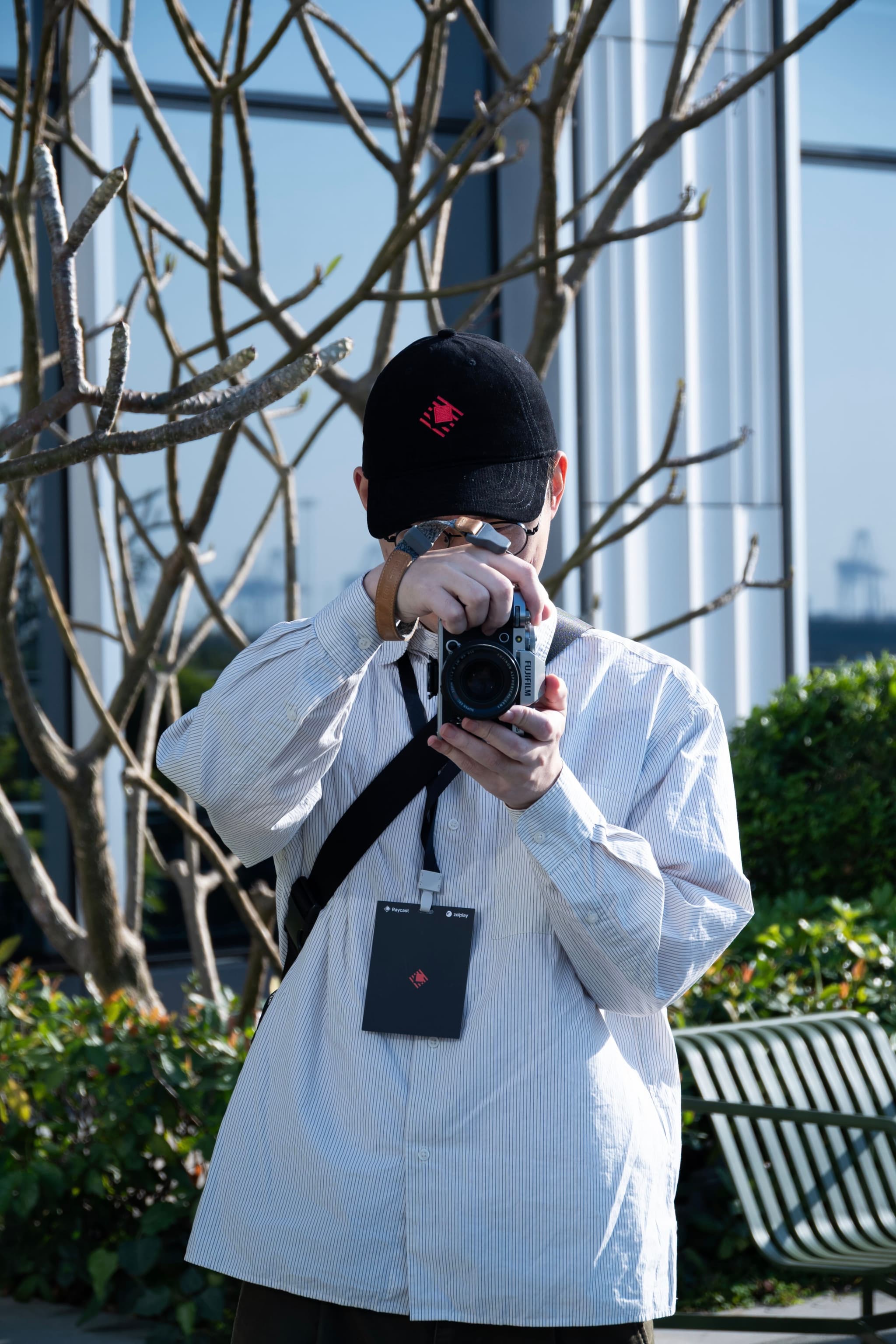 Person in a white jacket and black cap taking a photo with a camera near leafless trees and a bench in an urban park