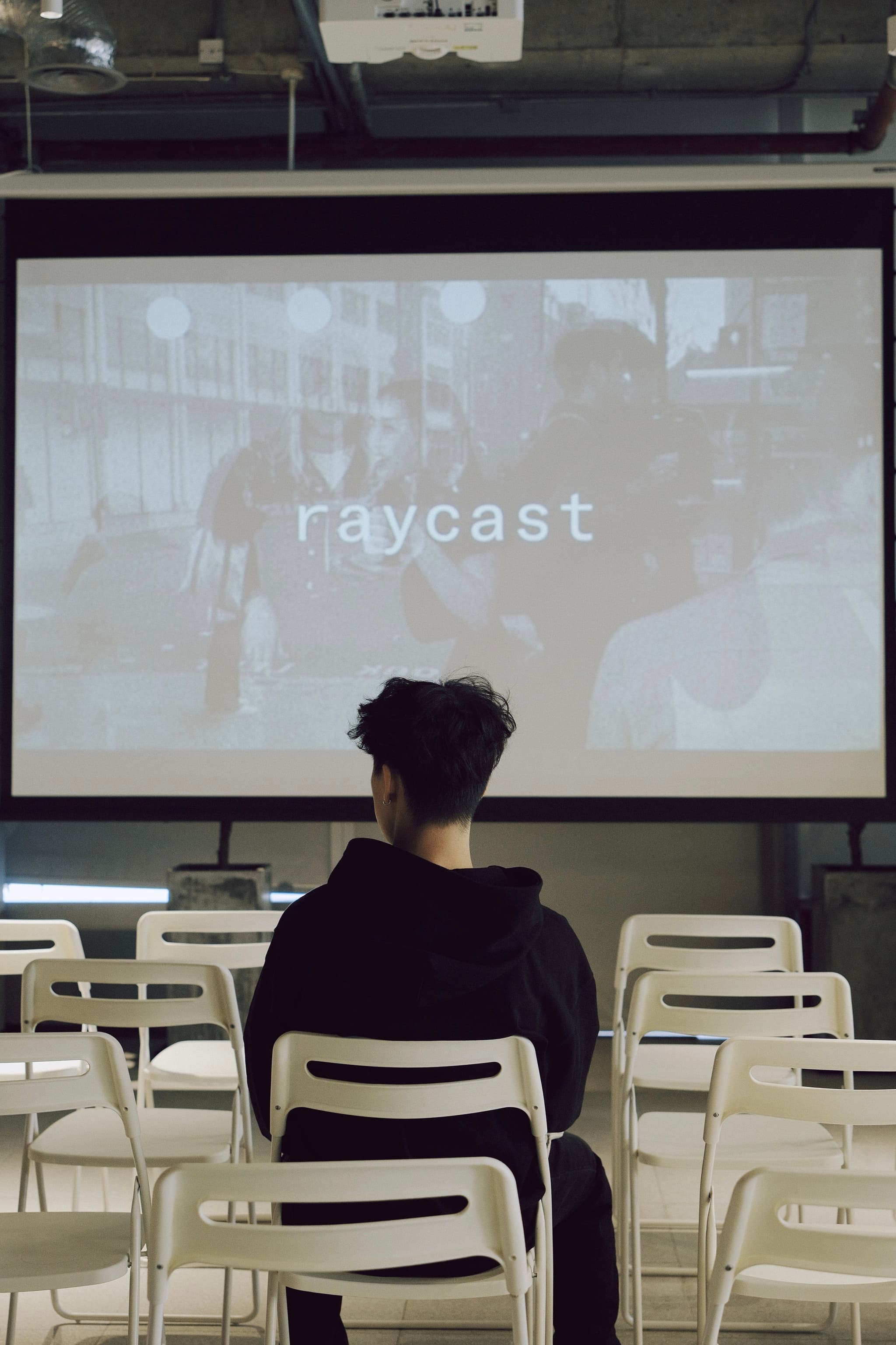 Person seated among empty white chairs facing a projector screen displaying the word raycast in an industrial indoor venue