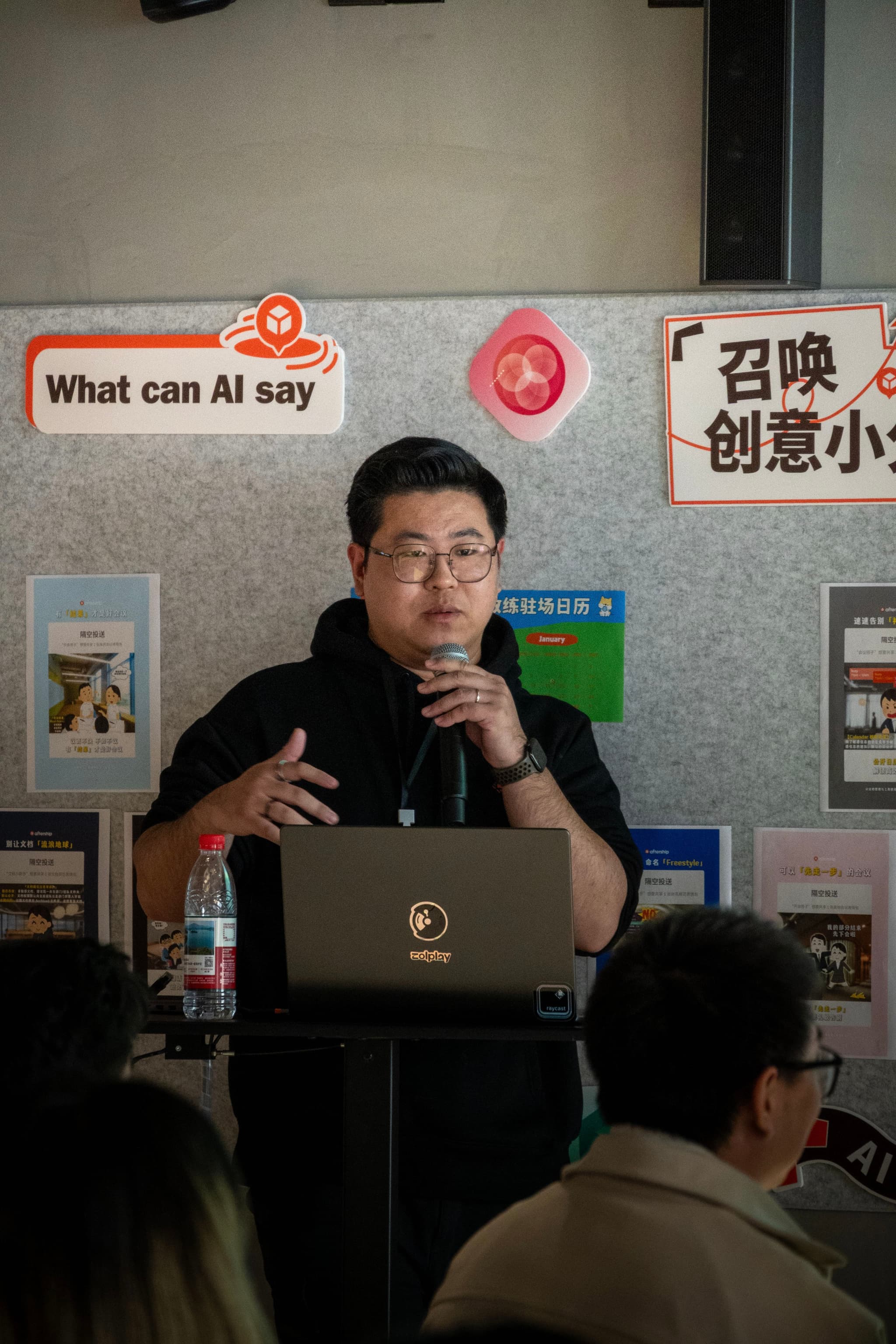 Man with microphone presenting behind laptop at small event with posters on wall