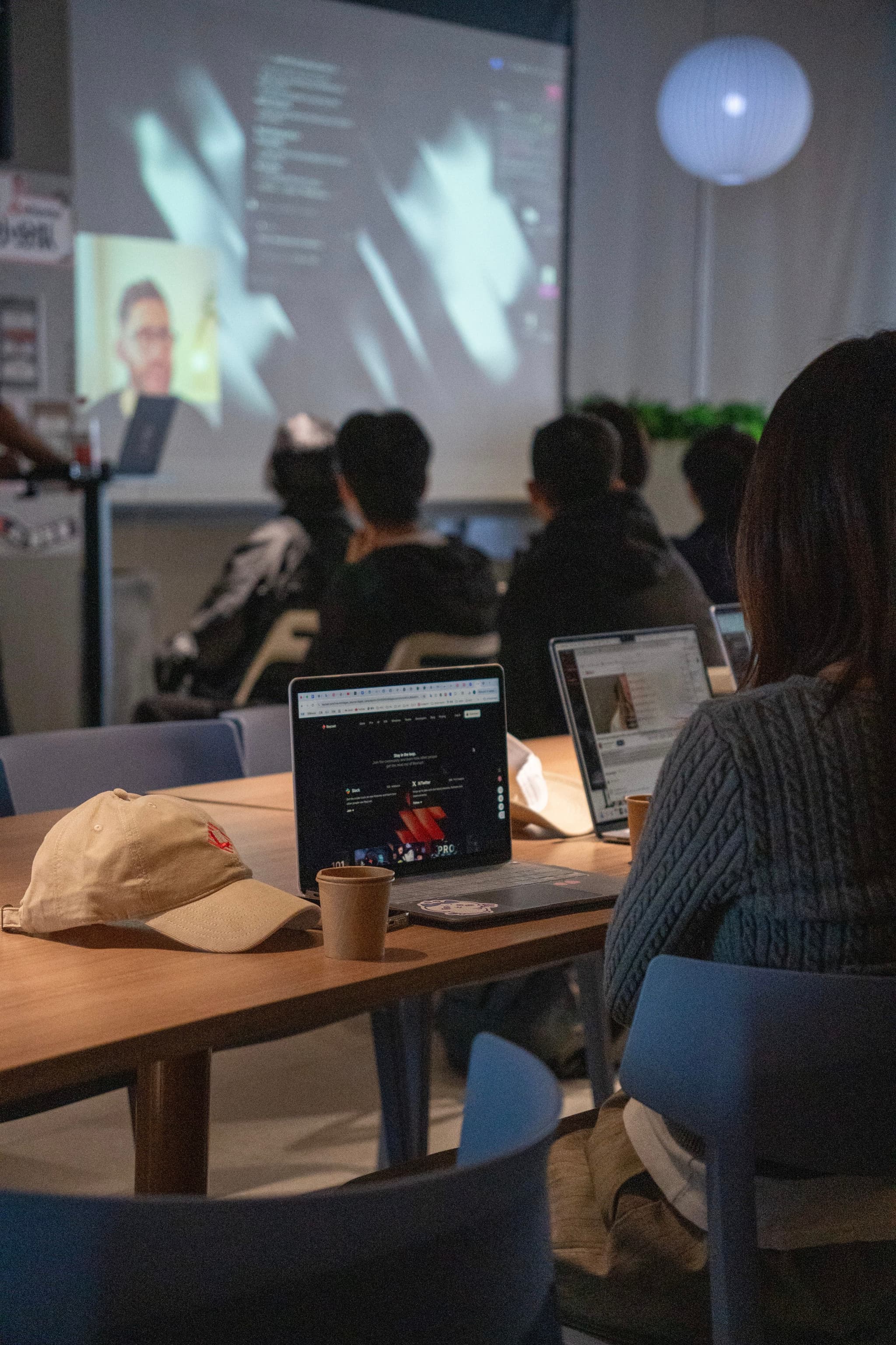 People seated at tables with laptops watching a projected presentation in a classroom setting