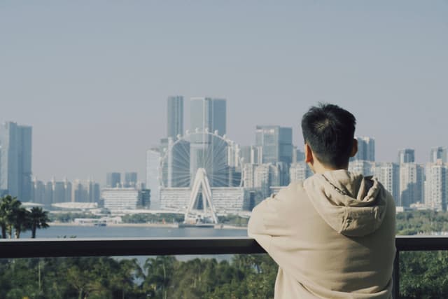 Person in a hoodie stands at a railing looking over a bay toward a modern city skyline with high rise buildings in hazy daylight