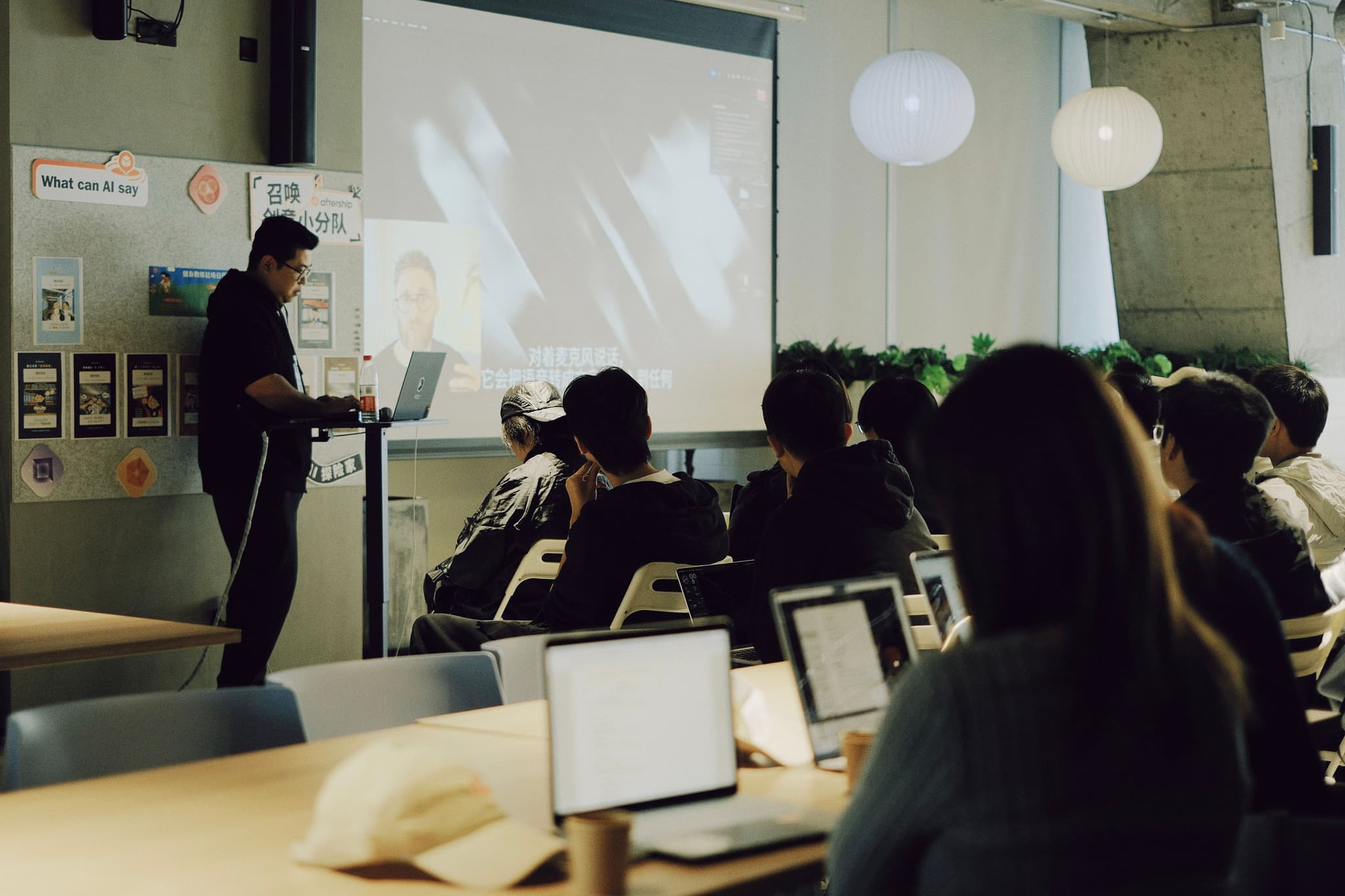 A person stands at the front of a classroom near a projection screen while students sit at desks with laptops watching a video presentation