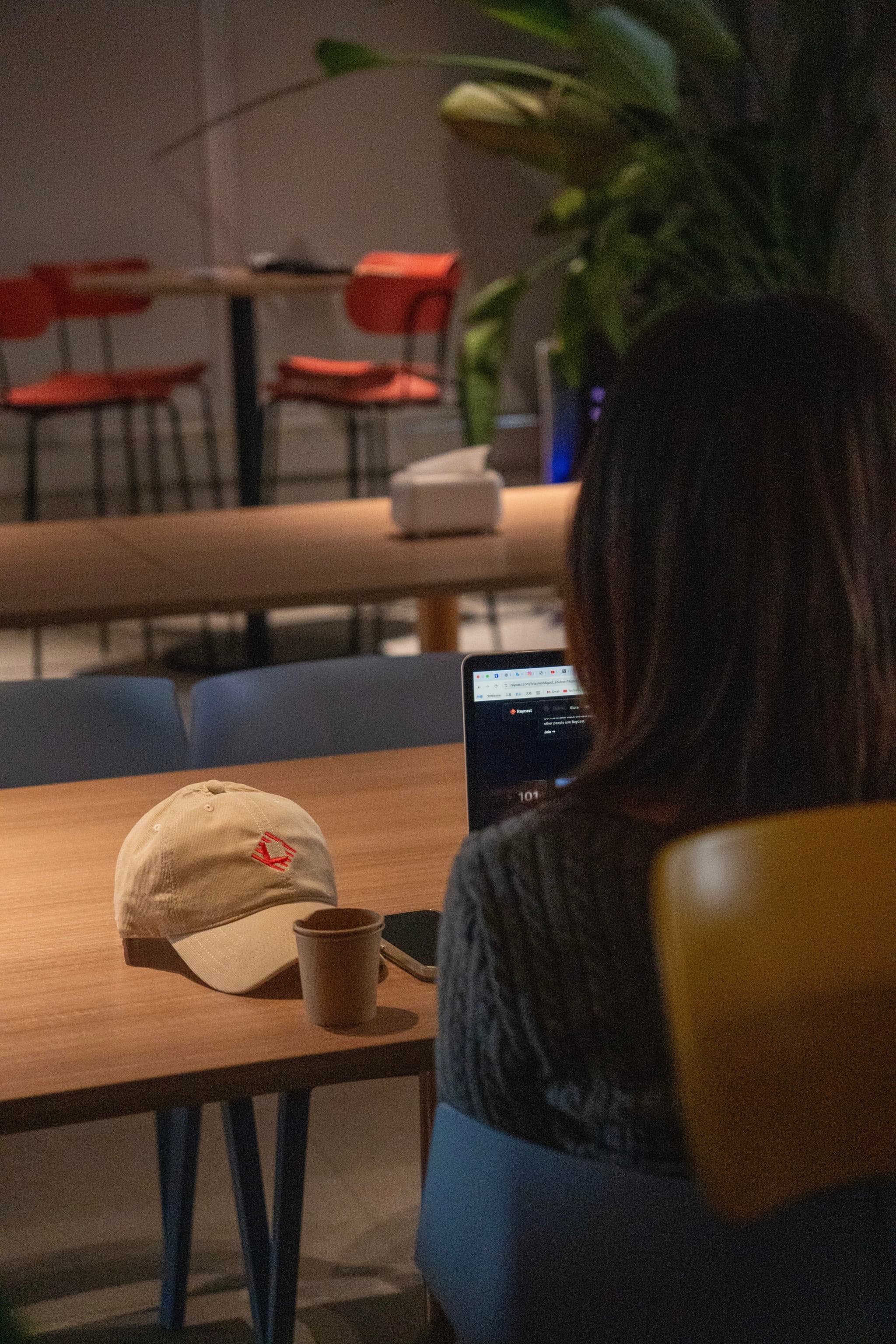 Person seated at a wooden table in a cafe working on a laptop with a cap and coffee cup nearby