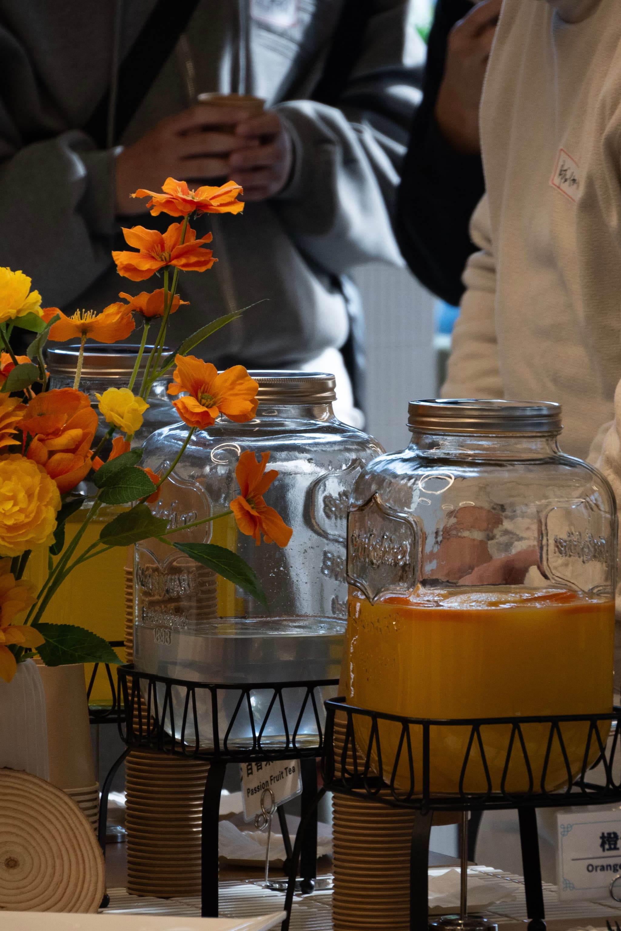 Glass beverage dispensers with orange juice on a table beside a small bouquet of orange and yellow flowers with people blurred in the background