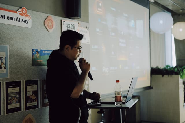 Person speaking into a microphone beside a laptop and projector screen in a small event space with posters on the wall