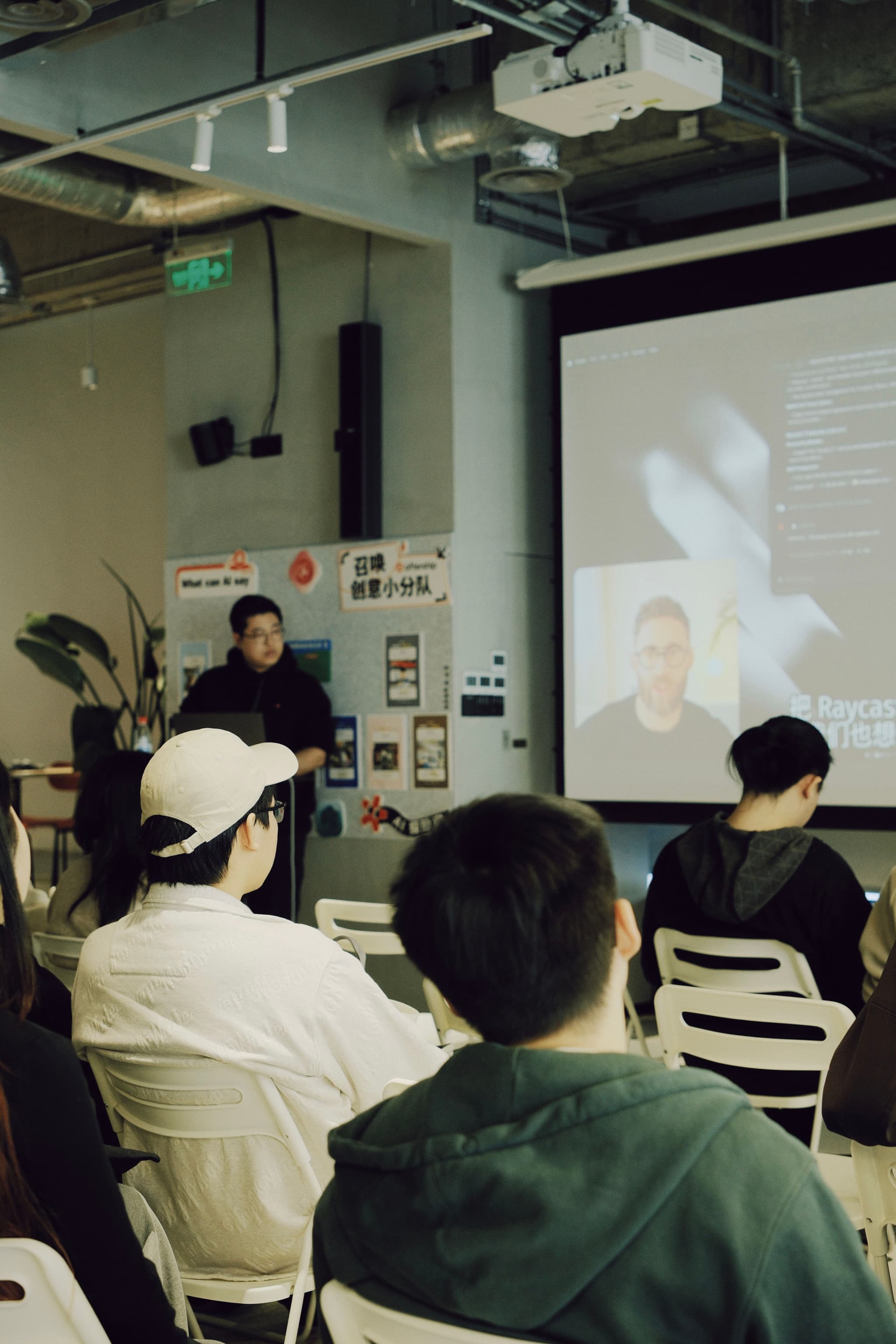 Small audience seated in a room watching a projected video while a presenter stands near the screen