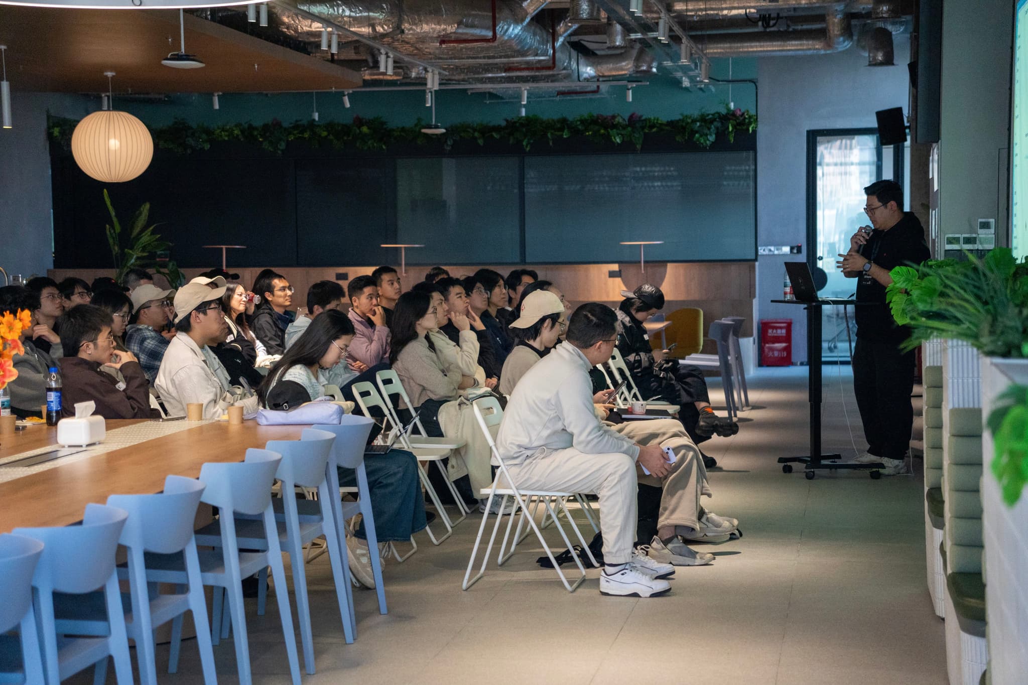 Group seated in a modern office cafeteria watching a standing presenter beside a screen