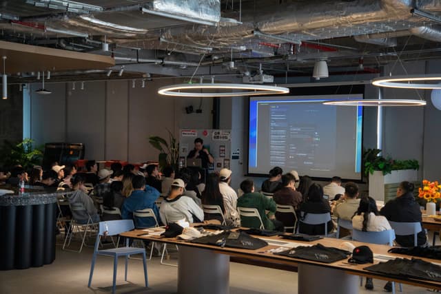 Open office event with seated attendees watching a projected presentation under industrial ceiling lights