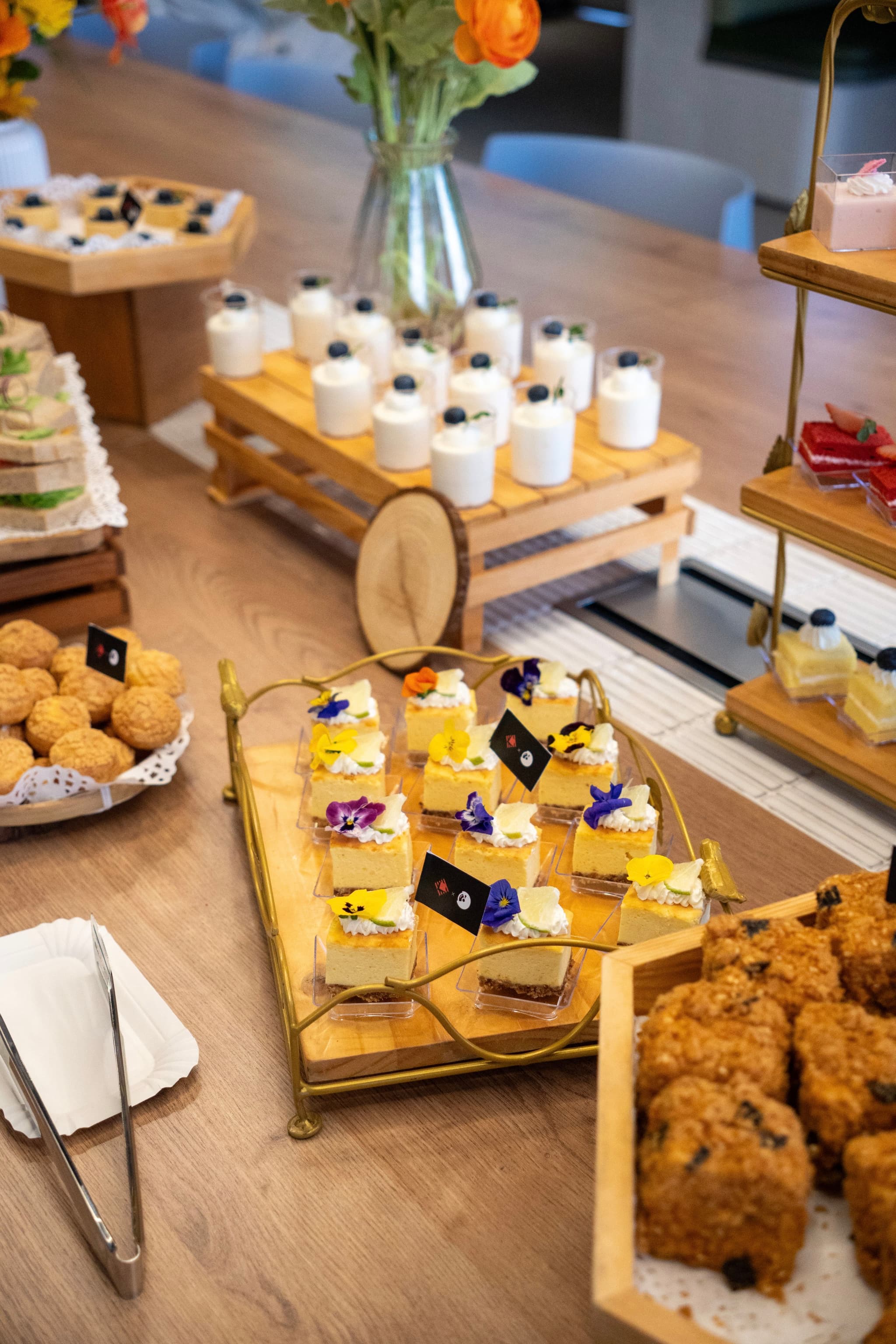 Assorted desserts and snacks on a buffet table with small labeled pastries drinks and flowers