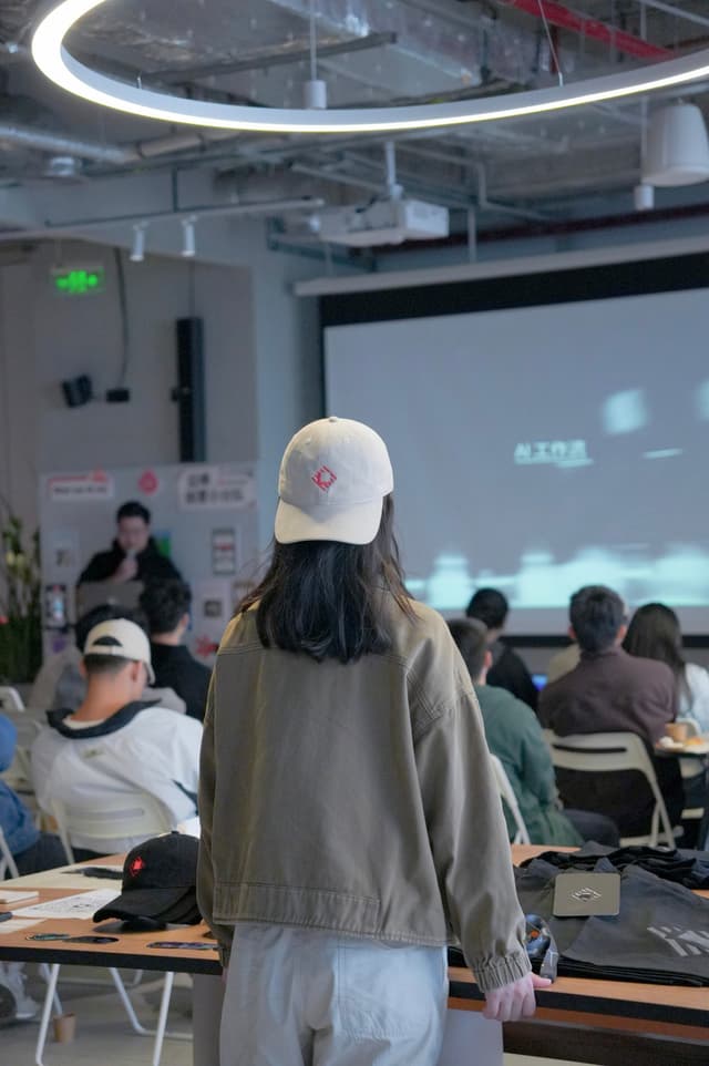 Student in cap standing in a modern classroom facing a projected screen while others sit at tables