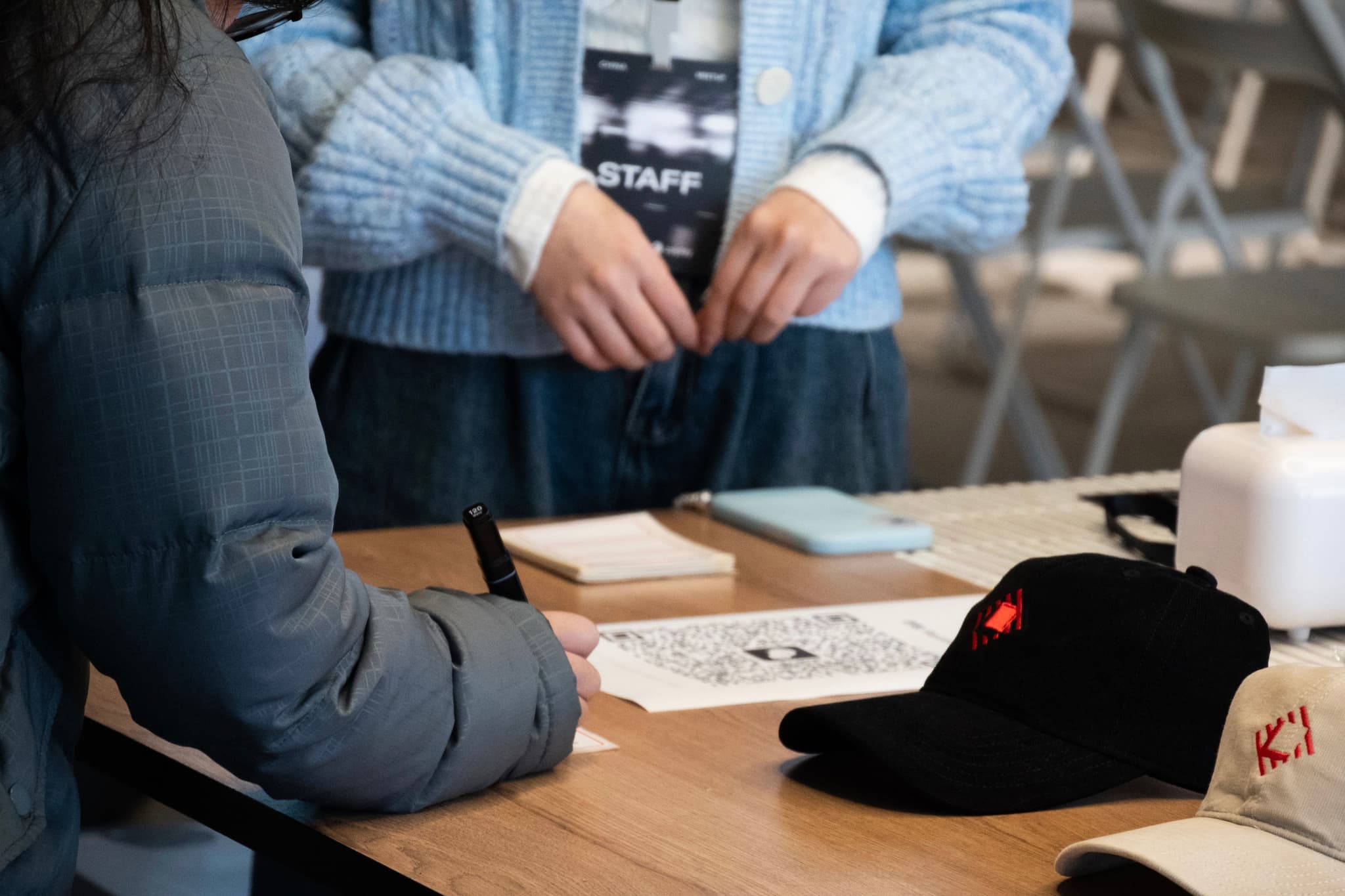Two people seated at a wooden table arranging tiles for a board game with cups and hats nearby