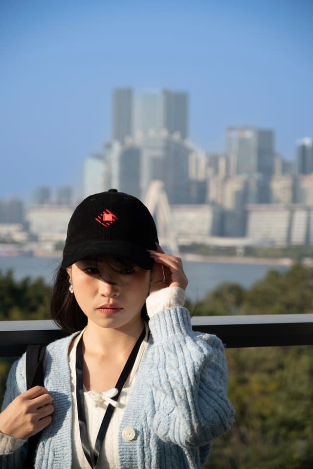 Young woman in cap and sweater on a balcony with a distant city skyline and water in the background