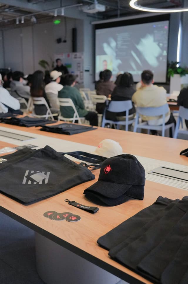 Conference room with attendees seated facing a projected presentation screen and tables in the foreground with branded tote bags and a baseball cap