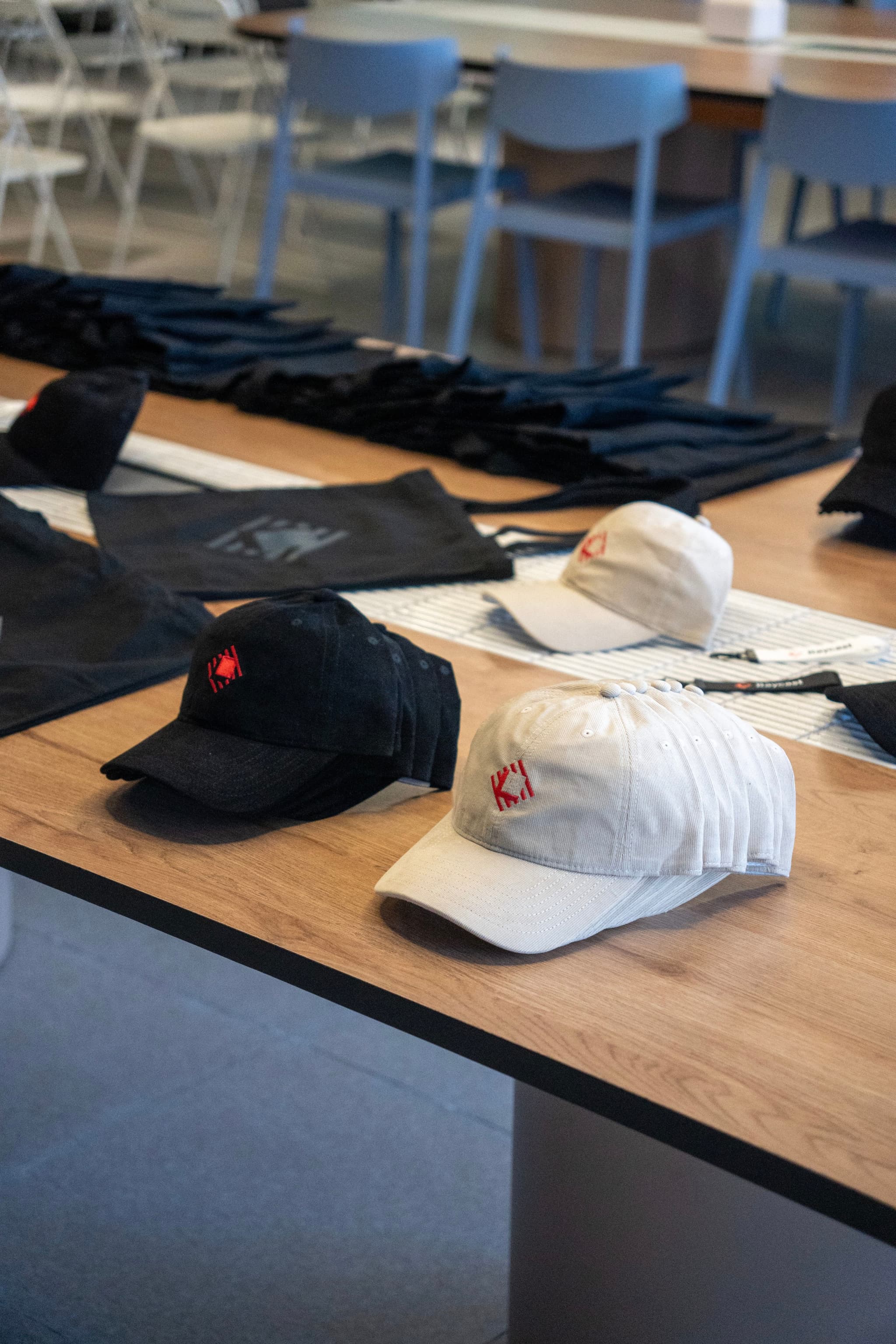 Assorted black and white branded baseball caps arranged on a wooden table in a room with chairs in the background