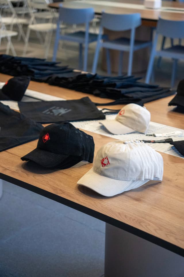 Assorted black and white branded baseball caps arranged on a wooden table in a room with chairs in the background