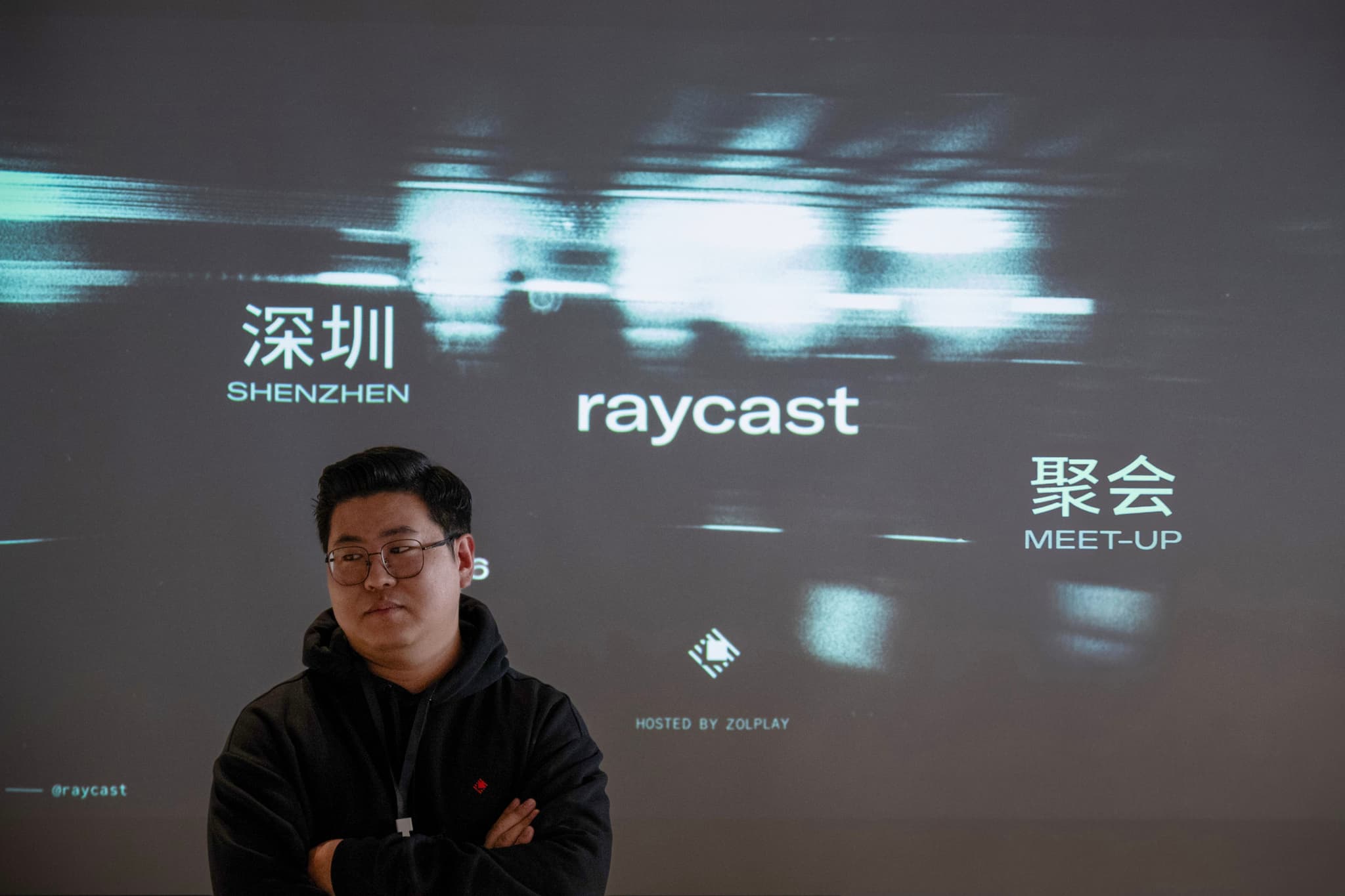 Person standing with arms crossed in front of a projected screen displaying Raycast branding and Shenzhen meetup text