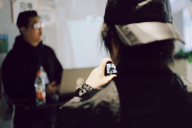 Person in cap and jacket filming with a handheld camera while another person stands in the background