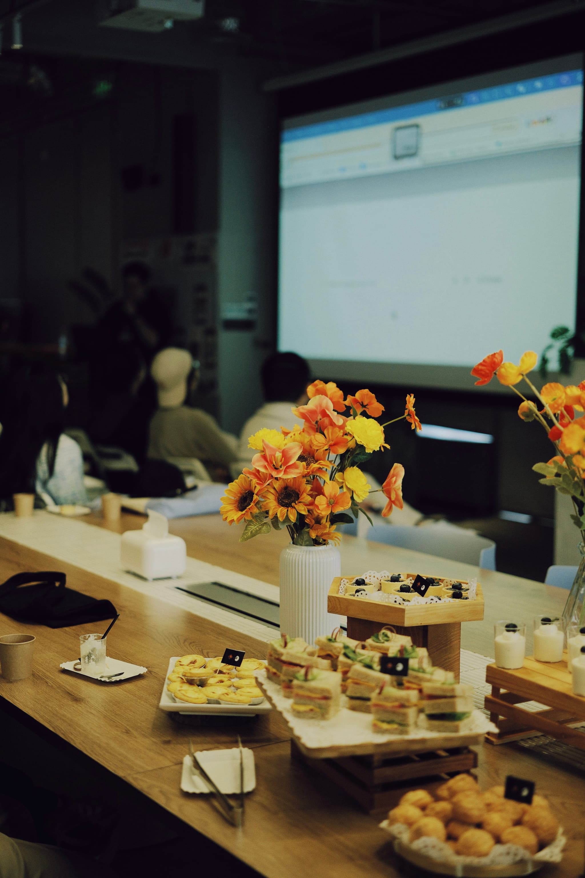 Conference room table with assorted snacks and orange flower arrangements facing a projected presentation screen