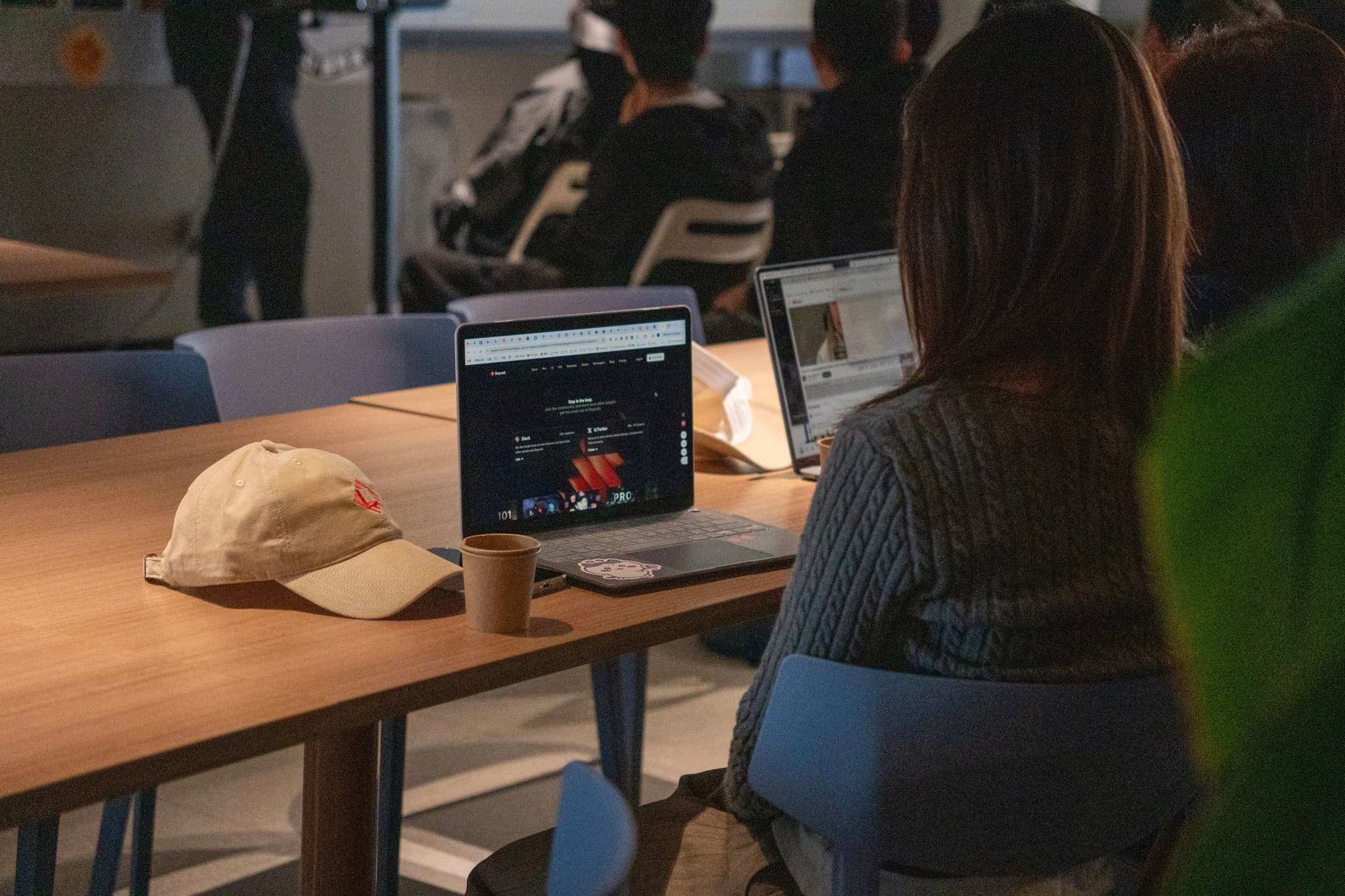 People seated around a table in a meeting space, watching video on a laptop with notebooks and a hat nearby