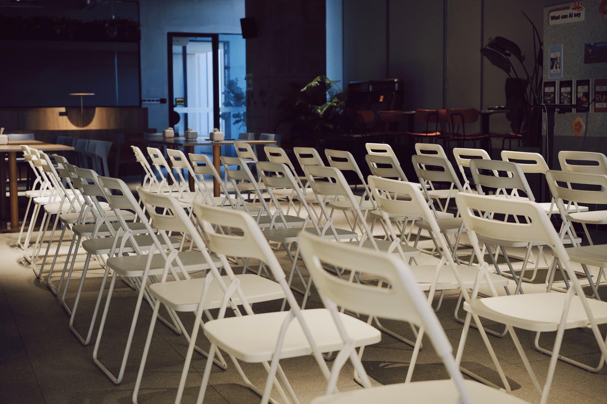 Rows of white folding chairs arranged in an empty indoor event room with equipment and tables in the background