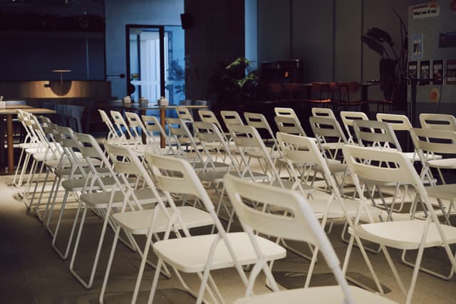 Rows of white folding chairs arranged in an empty indoor event room with equipment and tables in the background