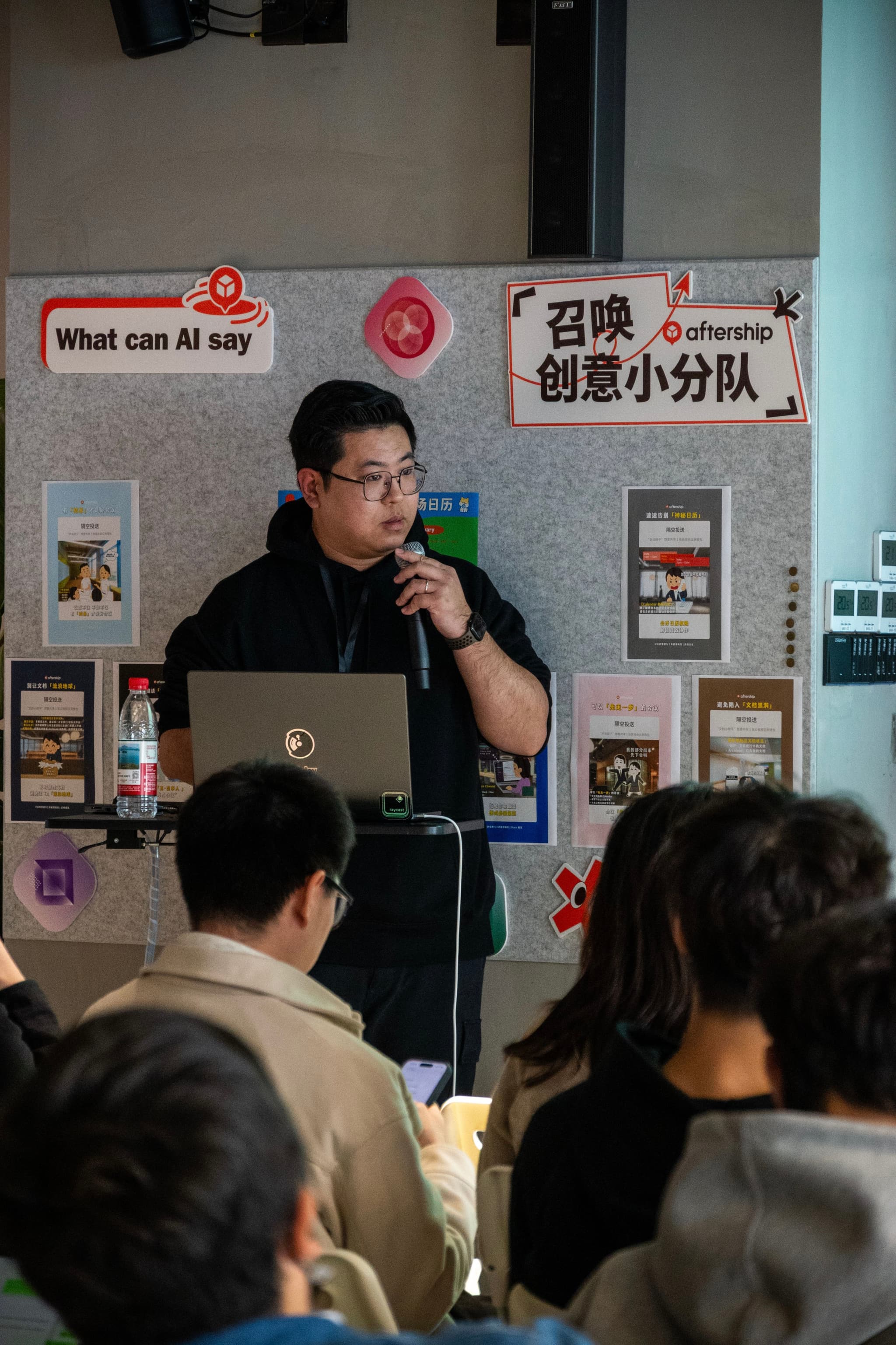 Man speaking into a microphone beside a laptop while an audience listens in a small indoor venue with posters on the wall