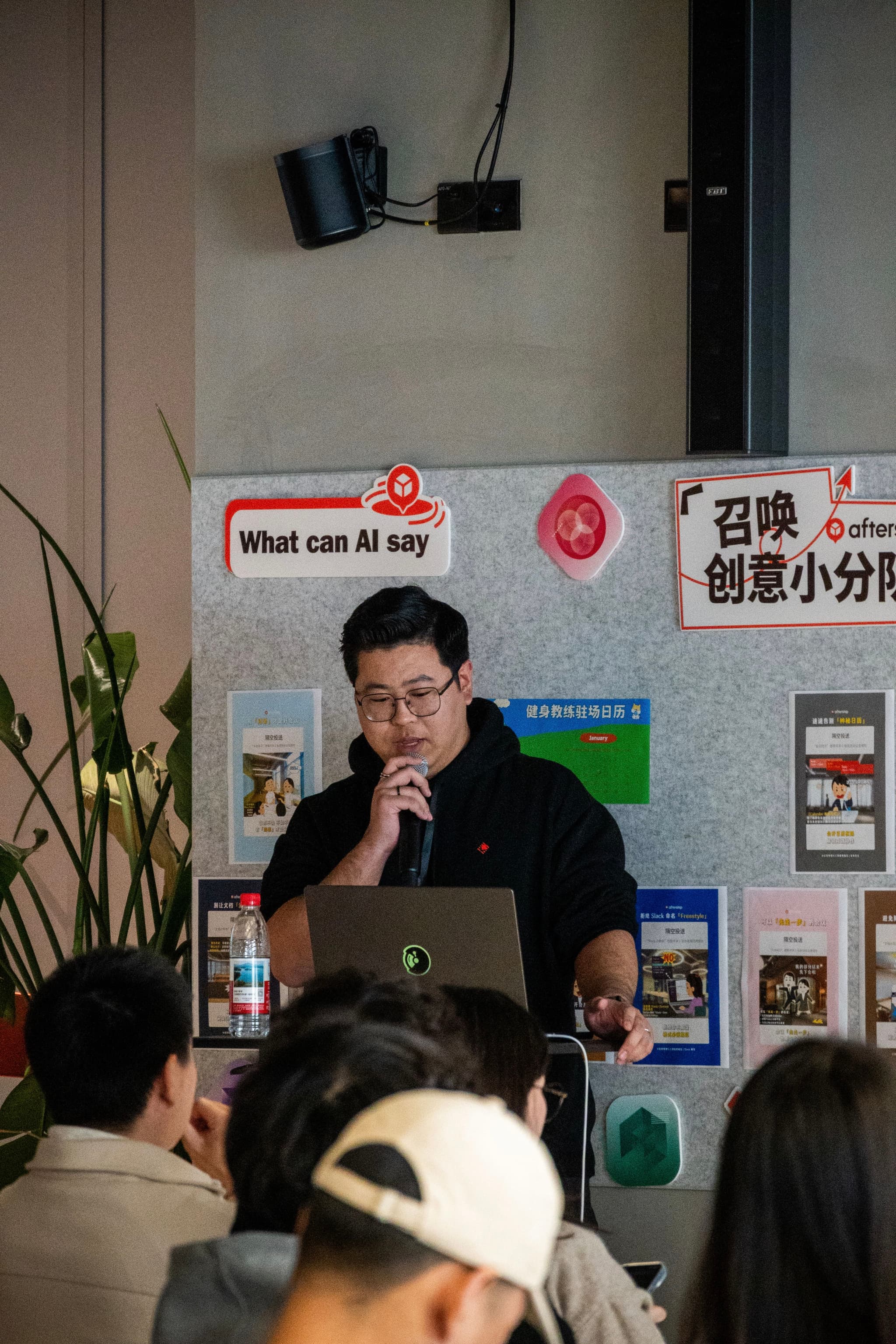 Man with microphone stands behind laptop speaking to seated audience in a small room with posters on the wall